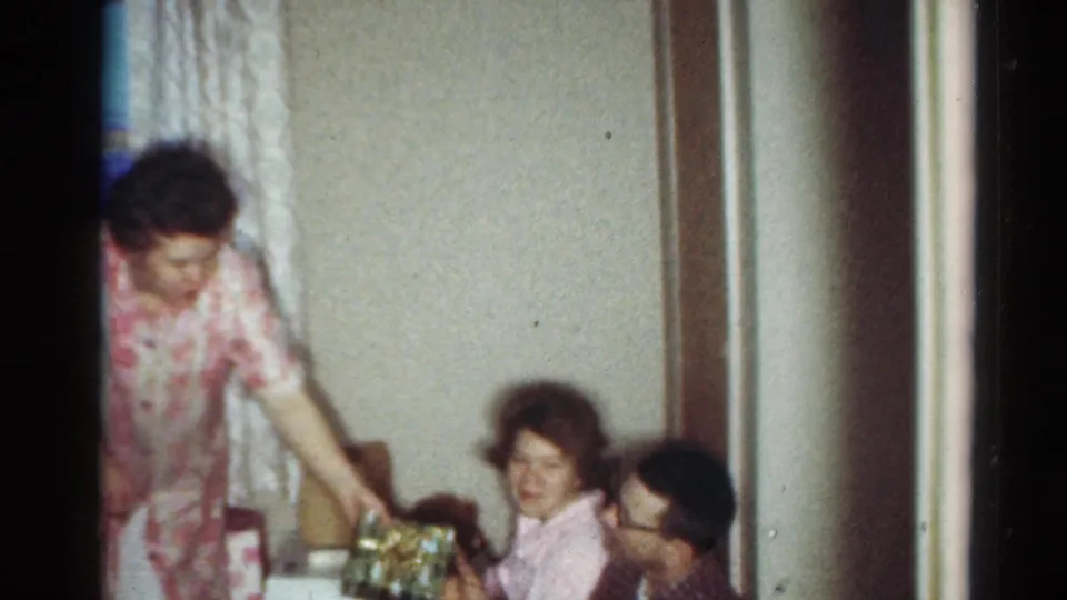 1964 Cleveland home-movie frame of a woman standing by seated relatives in a living room with wrapped gifts nearby.