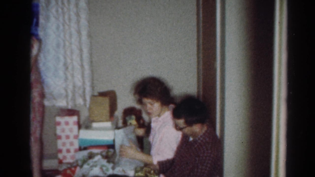 Seated woman and man look down at presents in a living room with gift boxes behind them.