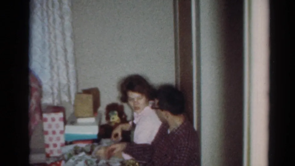 1964 Cleveland living-room frame with seated adults opening or sorting presents beside wrapped packages and curtains.