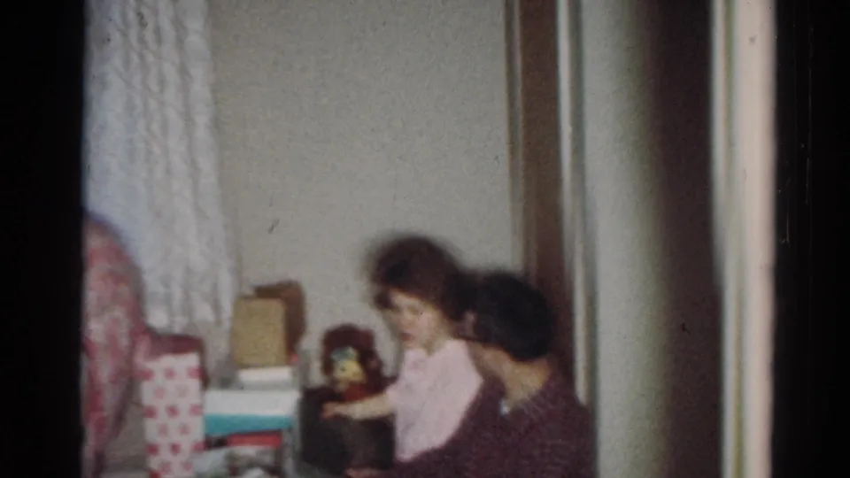1964 Cleveland home-movie frame of seated relatives beside wrapped gifts, curtains, and a small toy or doll in a private interior.