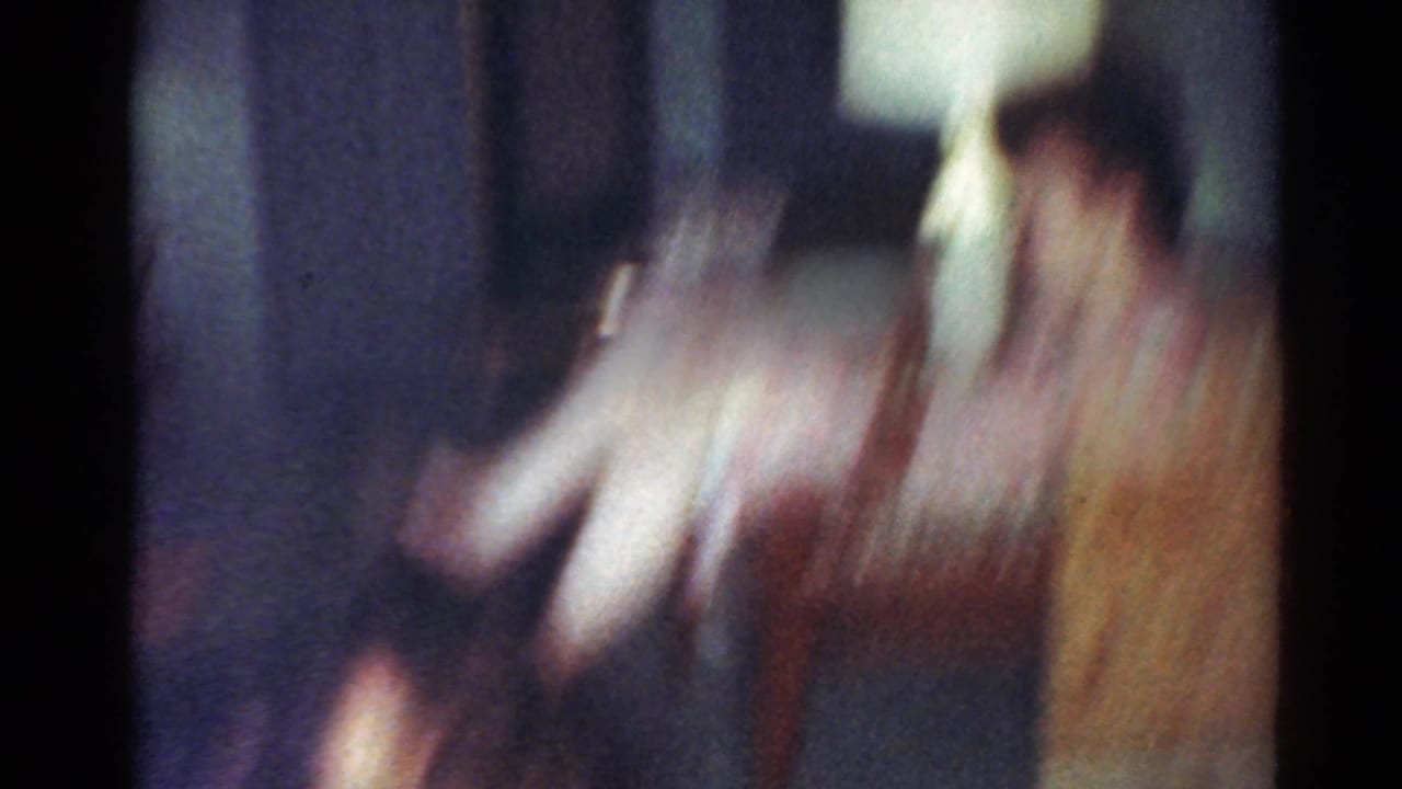 Soft, blurred 8mm frame from a 1964 Cleveland home movie showing figures moving around a dim dining area.