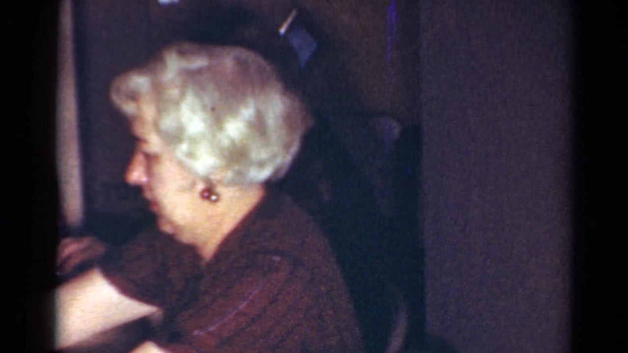 Older woman with styled white hair and earrings sits in profile in a dim Cleveland home interior, 1964.
