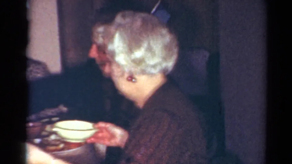 Older woman in a dark sweater holds a bowl at a dining table inside a 1964 Cleveland home-movie scene.