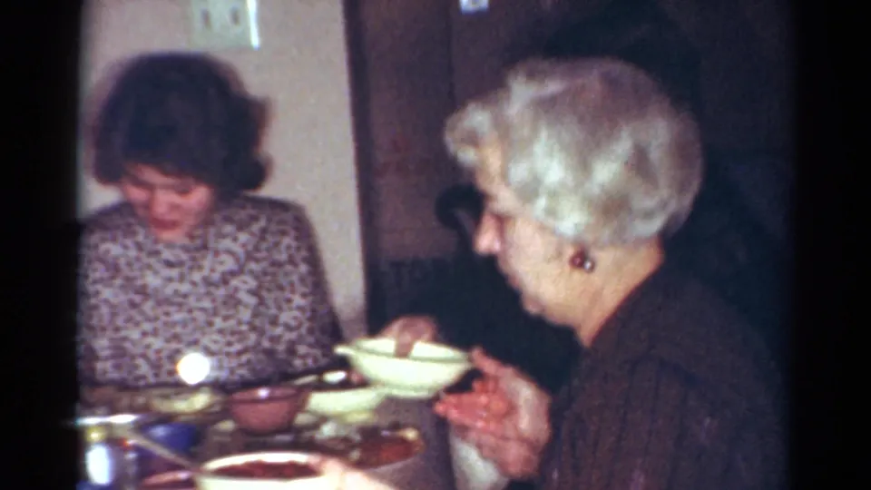 Two women sit at a dining table with bowls and dishes in a 1964 Cleveland 8mm home-movie dinner scene.