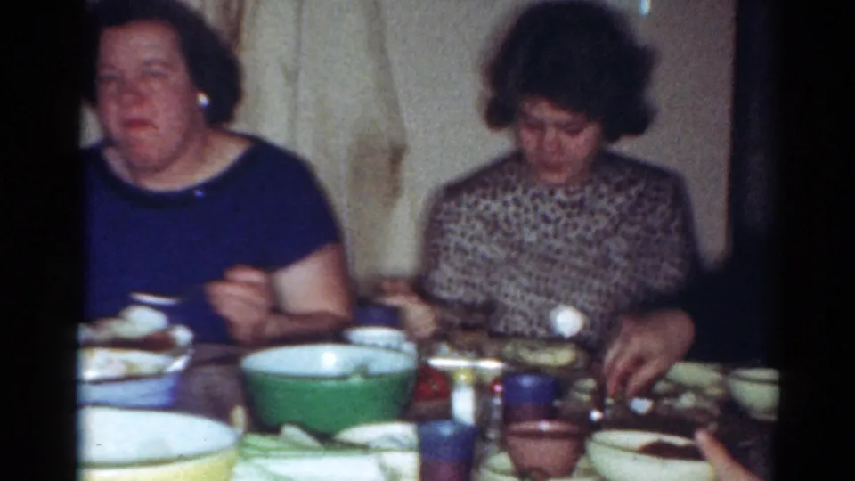 Adult women sit side by side at a 1964 Cleveland dinner table, eating from plates and bowls in a softly lit room.