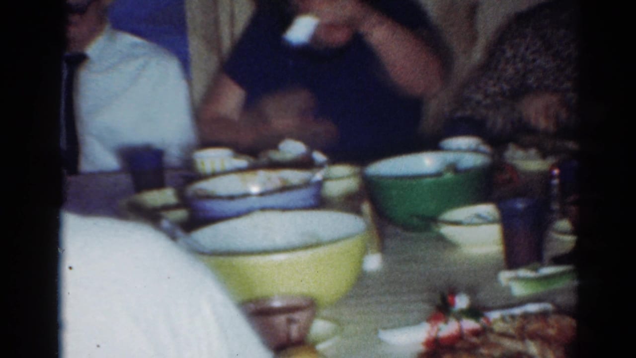 Cleveland 1964 family dining scene with serving bowls, cups, and adults partially framed by the handheld camera.