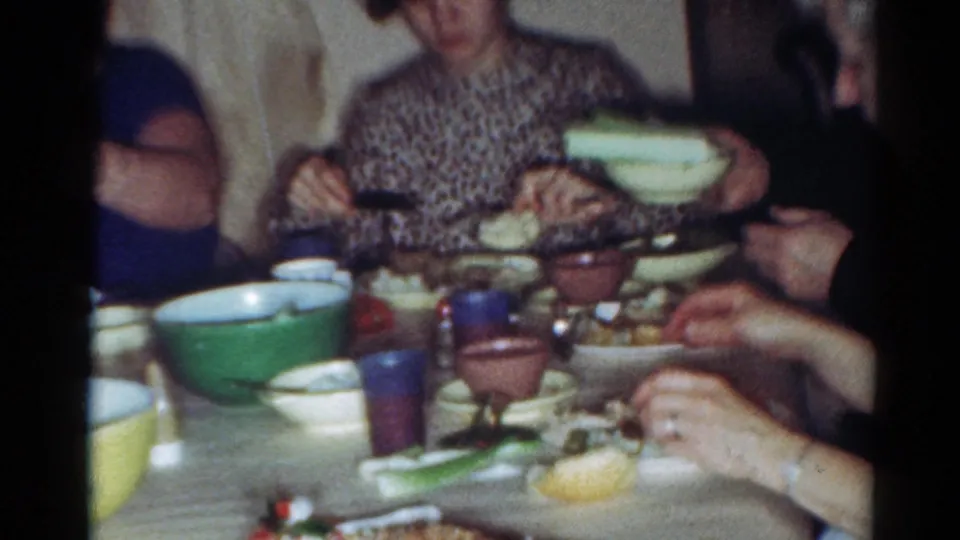 Cleveland 1964 home-movie frame of a woman in a patterned dress at a family dinner table with serving bowls and cups.