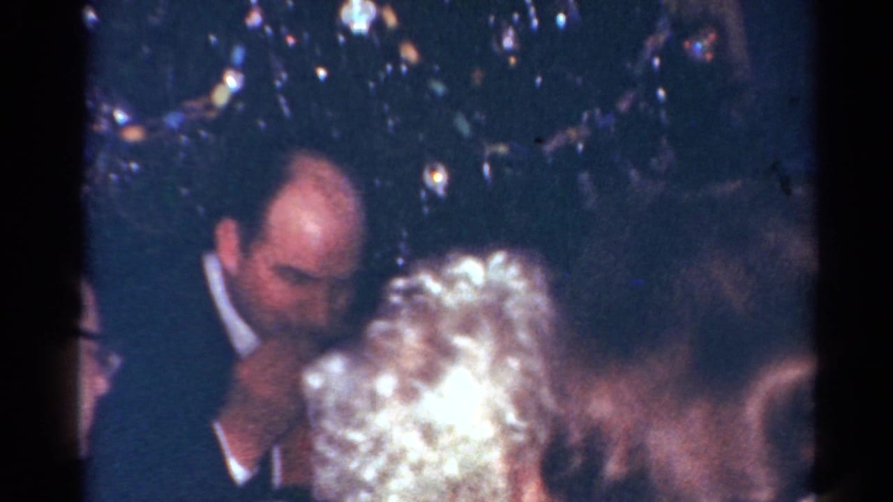 1964 Cleveland home-movie frame of adults seated tightly around a Christmas dinner table near tree decor.