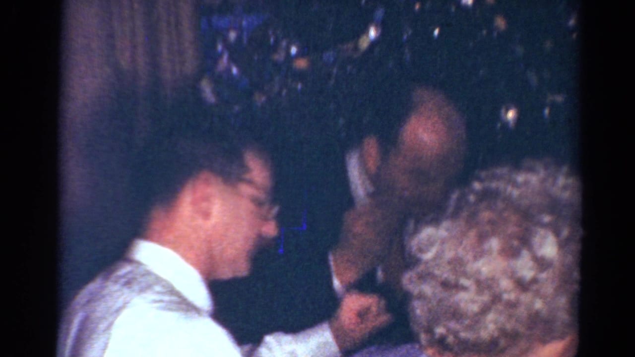 Adults in suits and light shirts gather near a table during a 1964 Cleveland holiday interior scene.