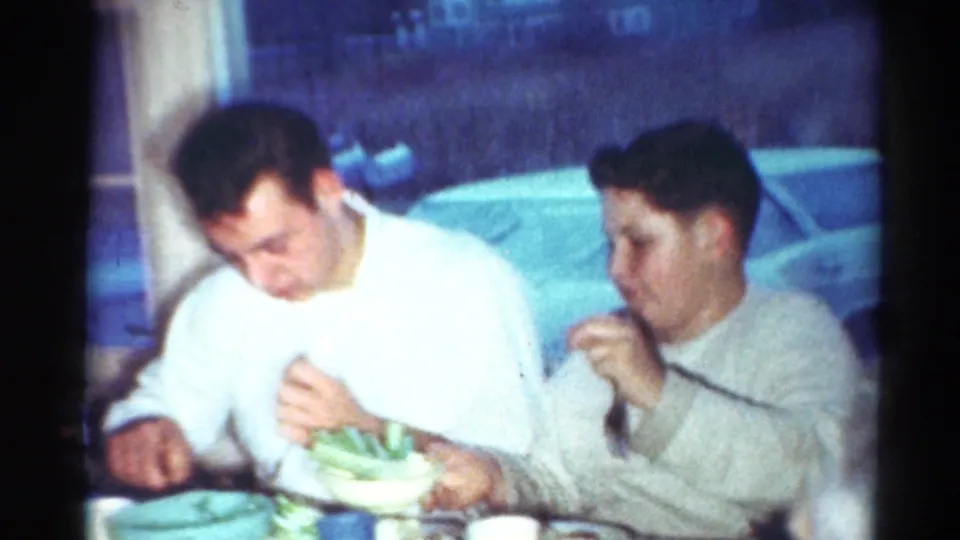 Two young men sit at a table in Cleveland, eating beside a window with a parked vintage car visible outside.