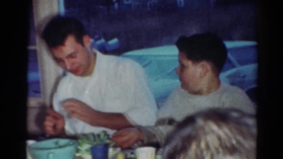 Two young men continue eating at a table in a 1964 Cleveland home interior, with dishes and a parked car visible through the window.