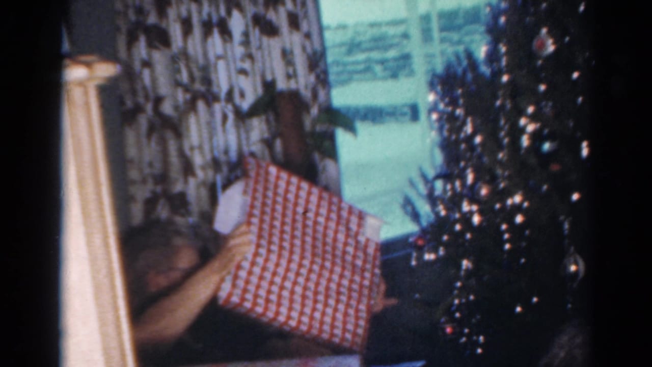 1964 Cleveland home-movie frame showing a figure with a wrapped present near a Christmas tree and patterned curtains.