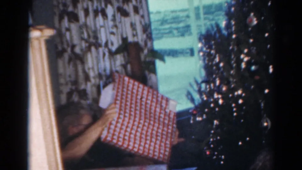 1964 Cleveland home-movie frame showing a figure with a wrapped present near a Christmas tree and patterned curtains.