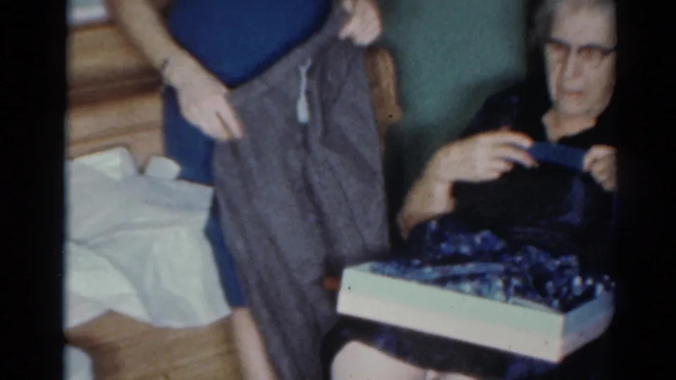 A woman in a blue dress holds up a gray garment for an older seated woman in a 1964 Cleveland home-movie interior.