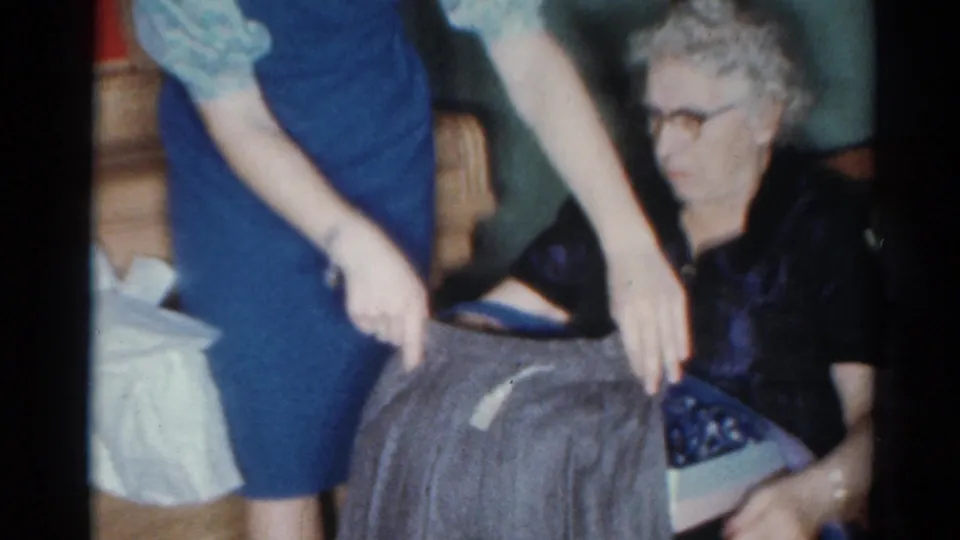The older woman in glasses looks toward the gray garment as it is held near a box in the 1964 Cleveland home interior.