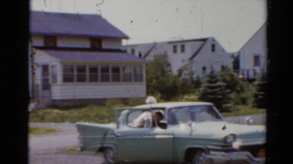 1964 Elkhart, Illinois home-movie frame showing a light green sedan stopped in a driveway near white suburban houses.