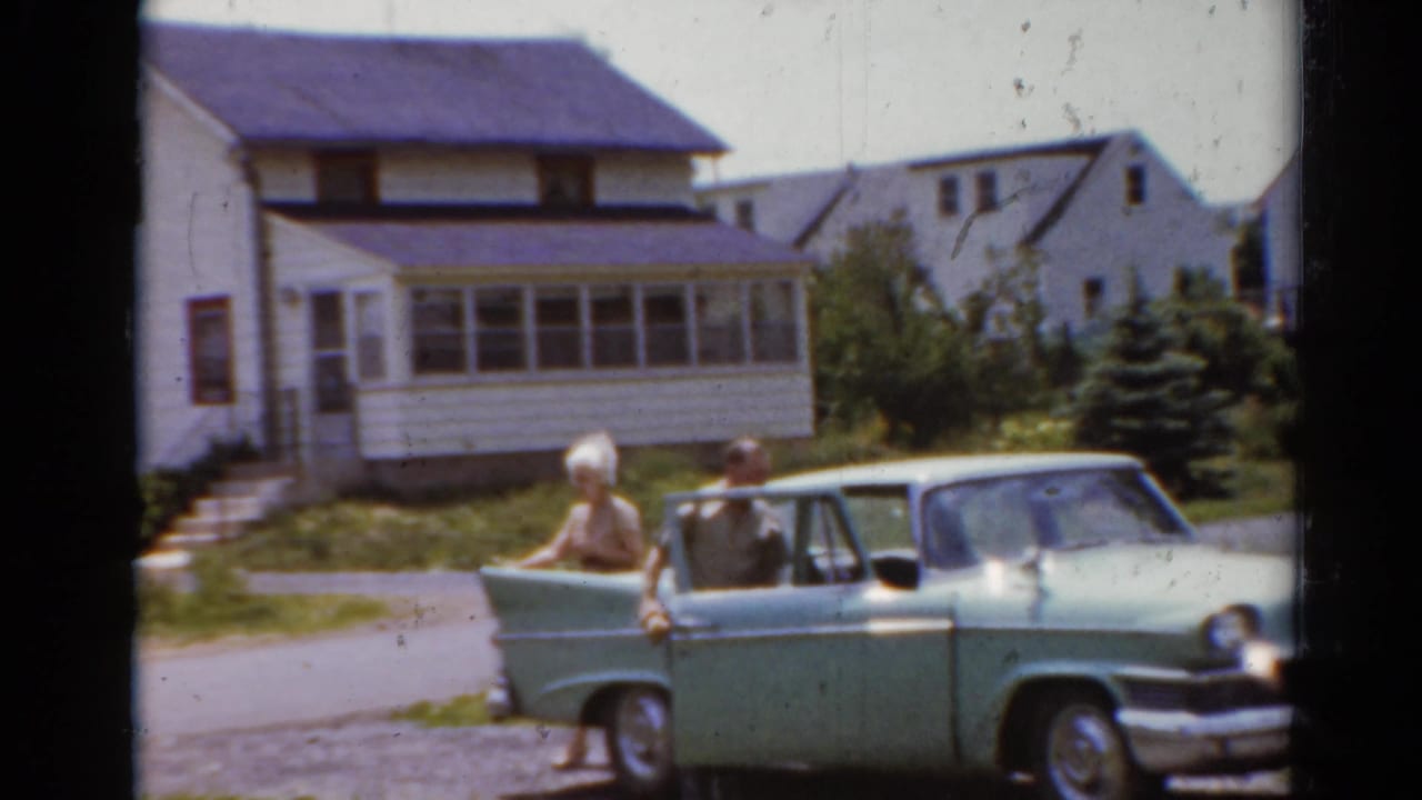 1964 Elkhart, Illinois frame with a woman beside the open rear door of a light green sedan in front of suburban houses.