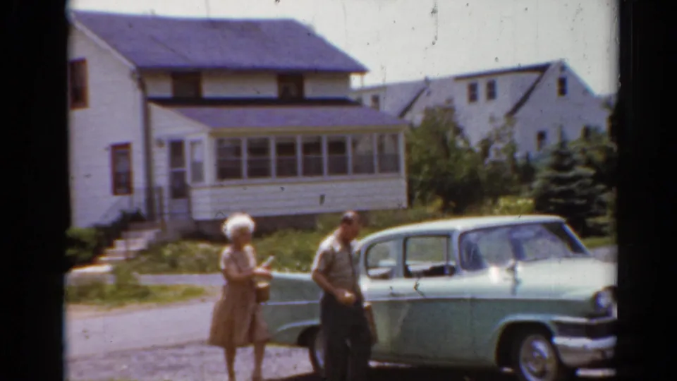 1964 Elkhart, Illinois frame with a man and woman near a light green sedan parked by white suburban houses.