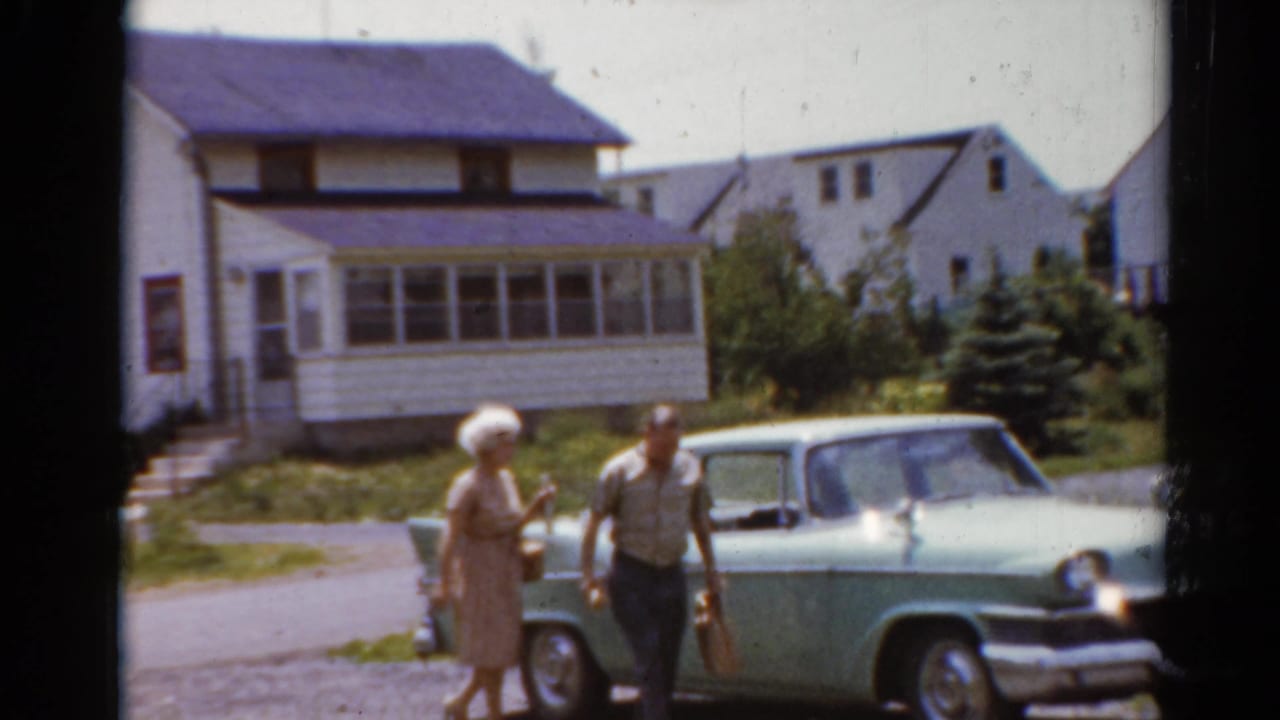 A woman and man stand beside a light green car in a residential neighborhood.