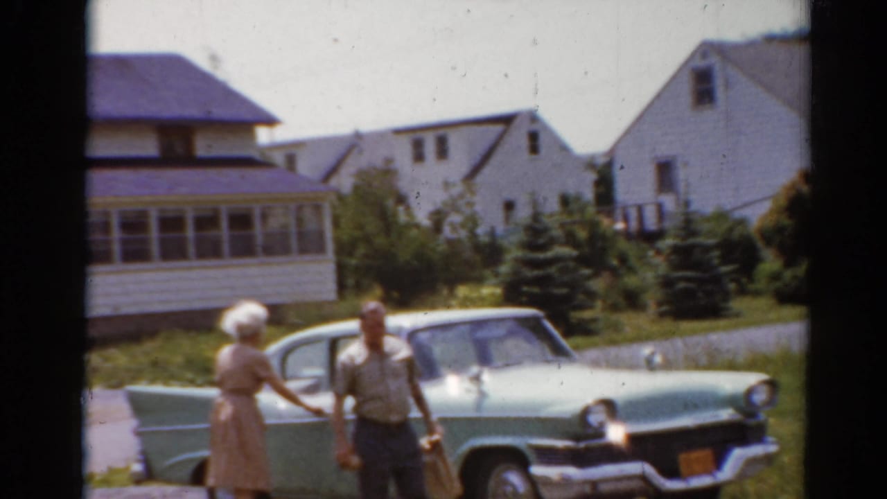 A woman reaches toward a man beside a light green car on a sunny street.