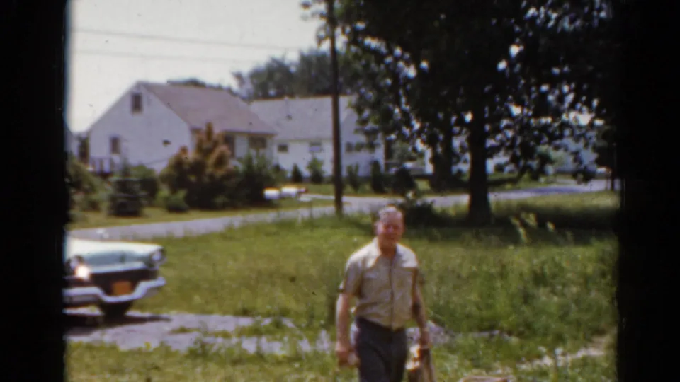 A man walks through a lawn in 1964 Elkhart, Illinois footage, with the car partly visible and residential houses in the background.