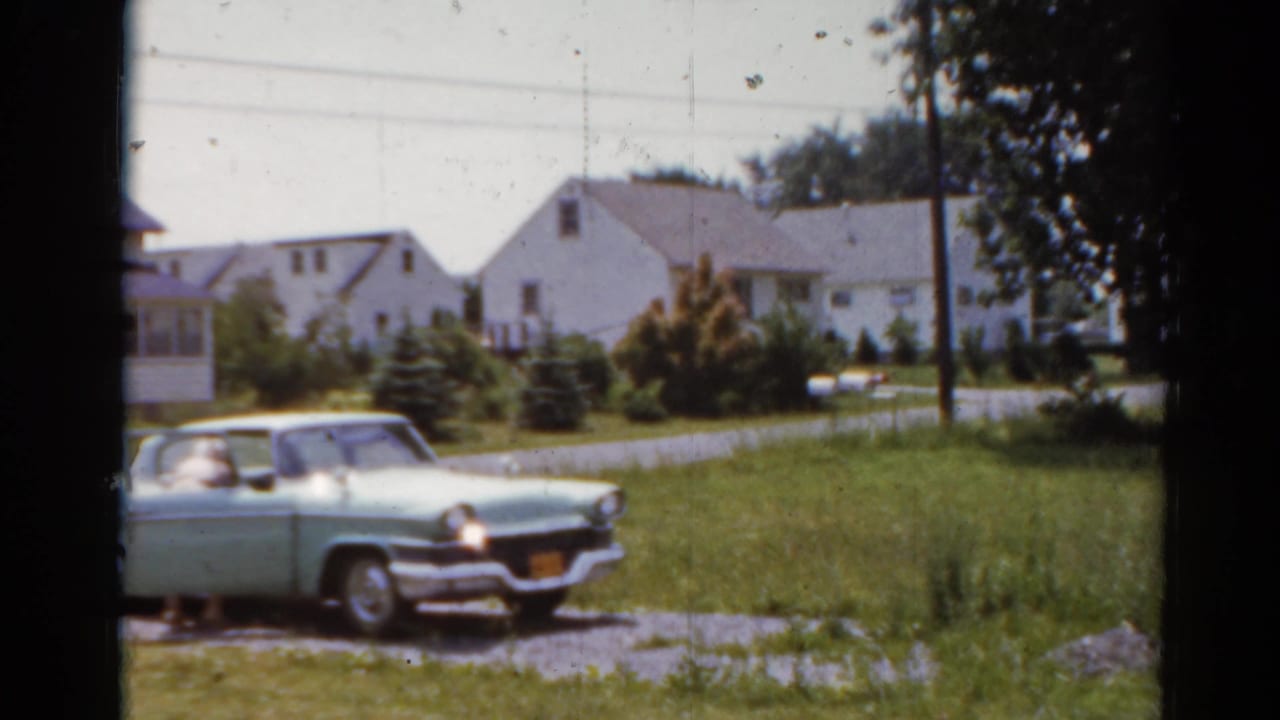 1964 Elkhart, Illinois home-movie frame showing a pale green car on a gravel drive near lawns and white houses.