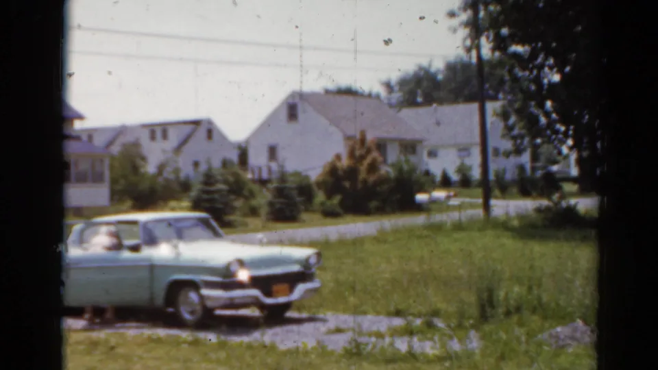 1964 Elkhart, Illinois home-movie frame showing a pale green car on a gravel drive near lawns and white houses.