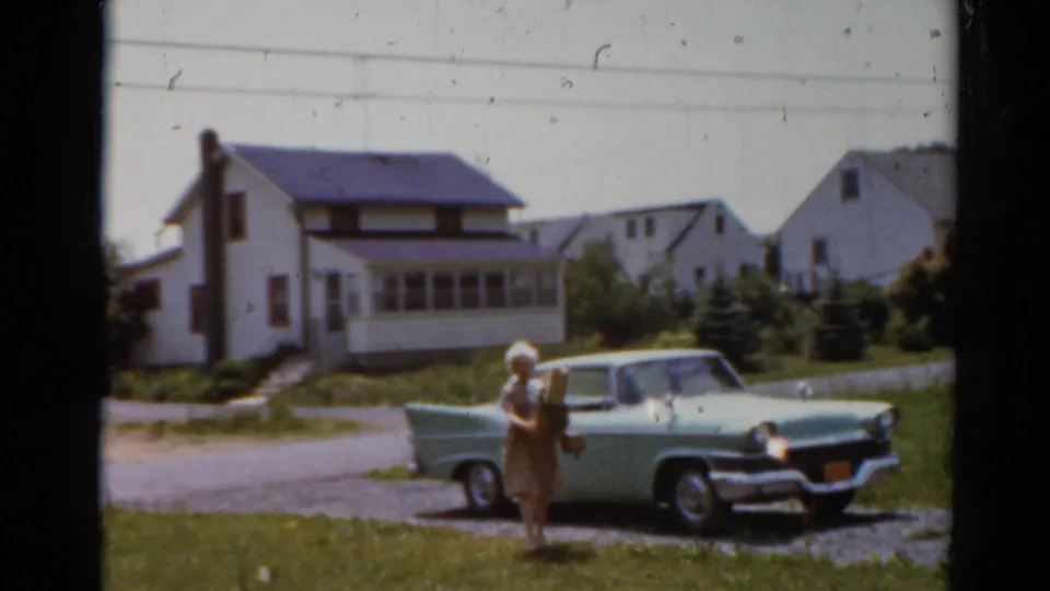 A woman holding a small child stands beside the pale green car in a 1964 Elkhart residential driveway scene.