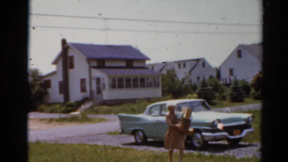 In Elkhart, Illinois in 1964, a woman carries groceries across a lawn near a pale green 1950s sedan and detached houses.