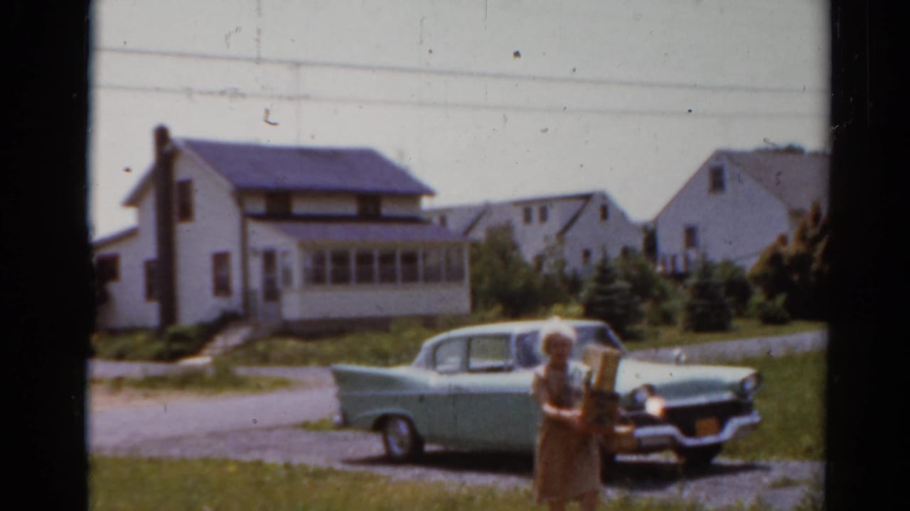 Woman with grocery bags stands closer to the camera beside a parked pale green sedan.