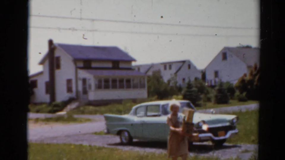 The woman carrying groceries moves nearer to the camera, with the 1950s sedan and Elkhart houses behind her.