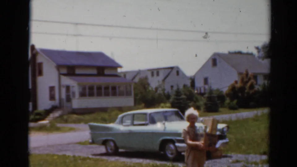 A 1964 home-movie frame shows the woman continuing across the lawn with groceries, with the parked sedan and houses behind.