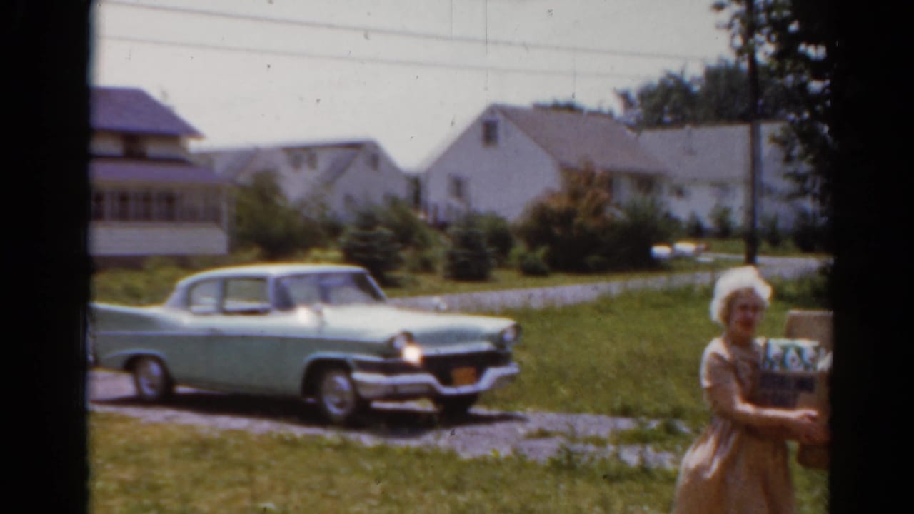 Woman carrying a box near a light green classic sedan in a suburban yard.
