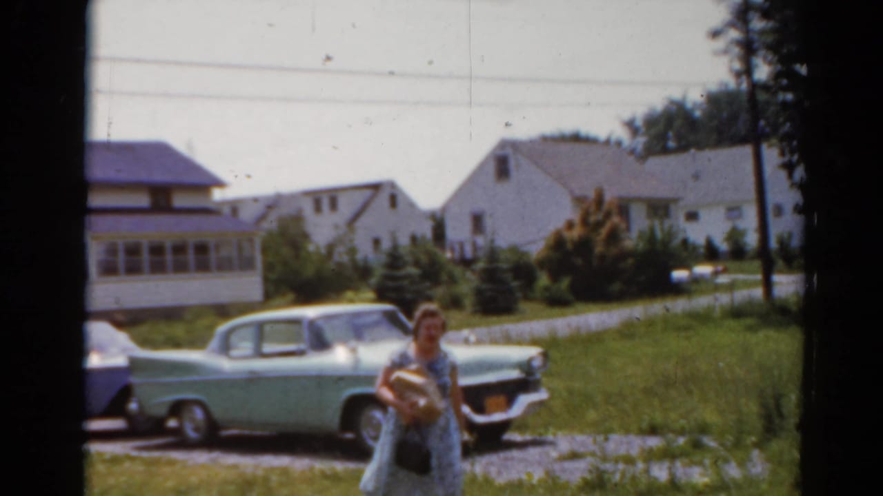 Person walking near parked classic cars in front of suburban houses.