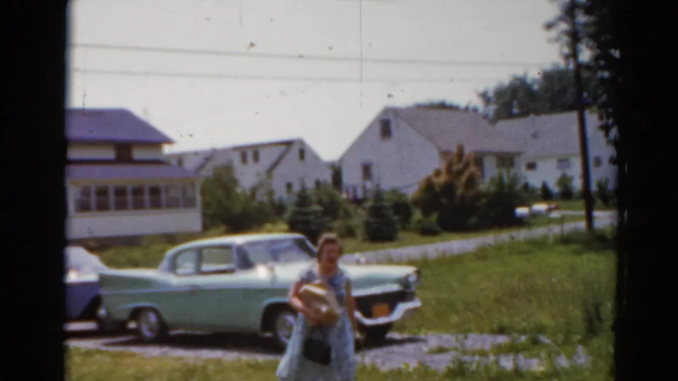 1964 Elkhart, Illinois frame showing a person walking by a light green sedan and blue car on a residential driveway.