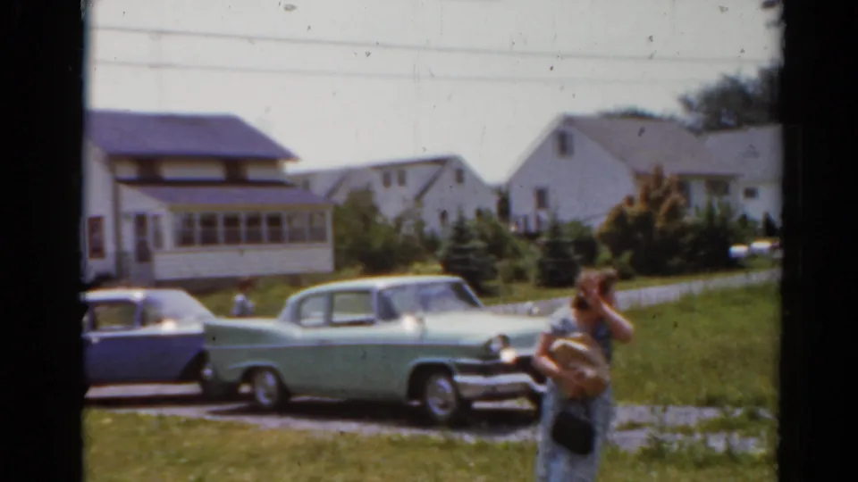 Home-movie frame from 1964 Elkhart, Illinois, with a person near two parked cars, a lawn, driveway, and white residential houses.