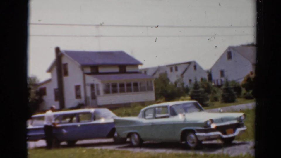 1964 Elkhart, Illinois frame showing two classic cars parked along a driveway before white houses and a grassy yard.