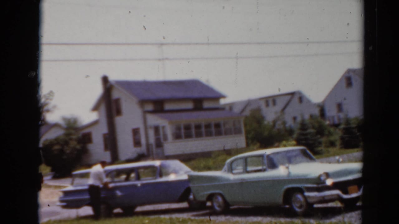 1964 8mm frame from Elkhart, Illinois showing a blue station wagon, a light green sedan, and a white suburban house.