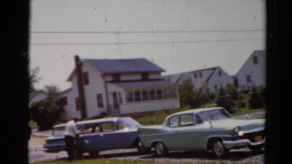 1964 8mm frame from Elkhart, Illinois showing a blue station wagon, a light green sedan, and a white suburban house.