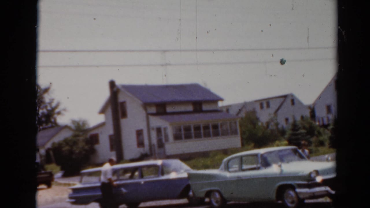 Elkhart, Illinois 1964 home-movie frame with parked classic cars, an adult figure, lawns, and neighboring houses.