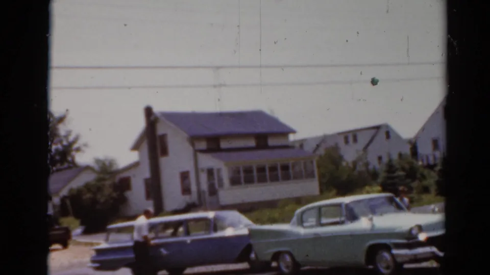 Elkhart, Illinois 1964 home-movie frame with parked classic cars, an adult figure, lawns, and neighboring houses.