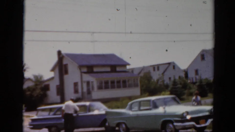 1964 Elkhart, Illinois 8mm frame showing an adult near a station wagon, a green sedan, lawns, and house exteriors.