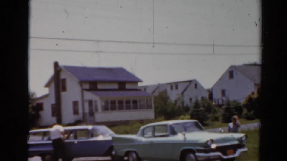 Elkhart, Illinois 1964 frame with classic cars, an adult standing by the station wagon, and suburban house exteriors.
