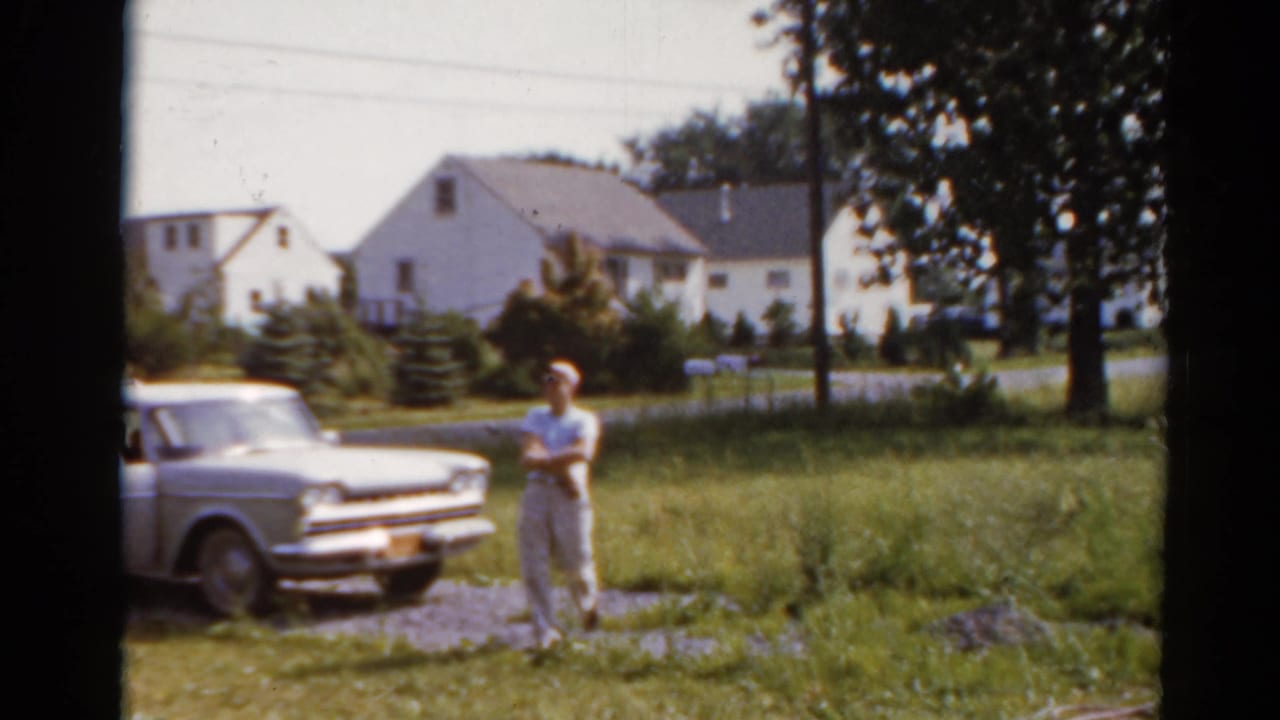 Boy stands near a light station wagon on a grassy residential street.