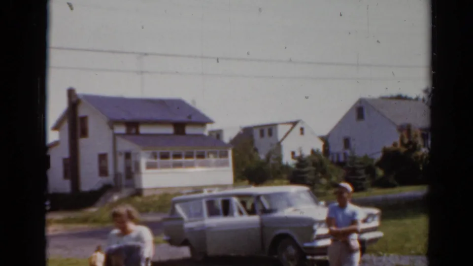 The woman with shopping bags appears close to camera while the station wagon, boy, houses, and lawns remain in the background.