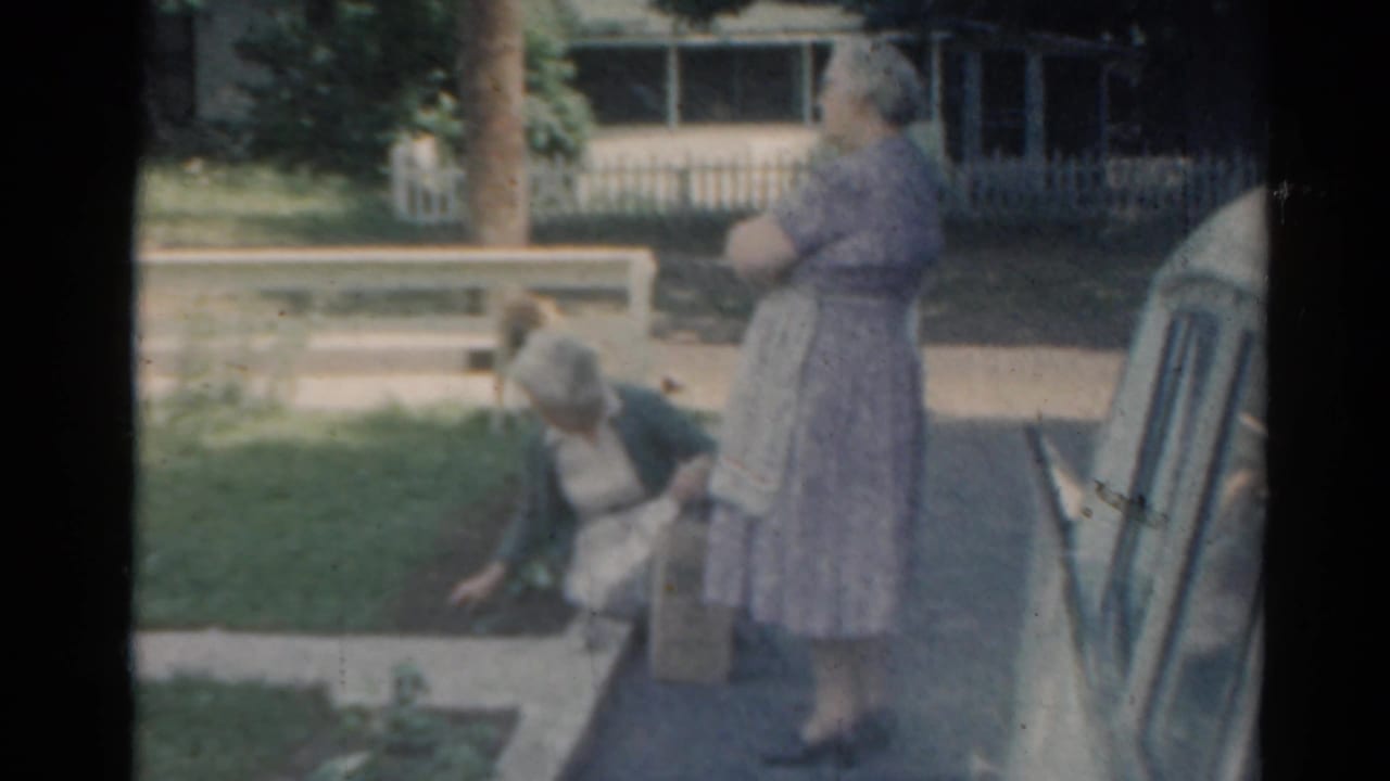 Two women in a suburban yard, one kneeling by a garden bed and one standing near a parked car.