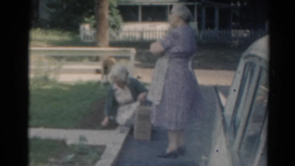 In this 1964 home-movie frame, two women are beside a suburban garden bed, with a parked car and white fence visible behind them.