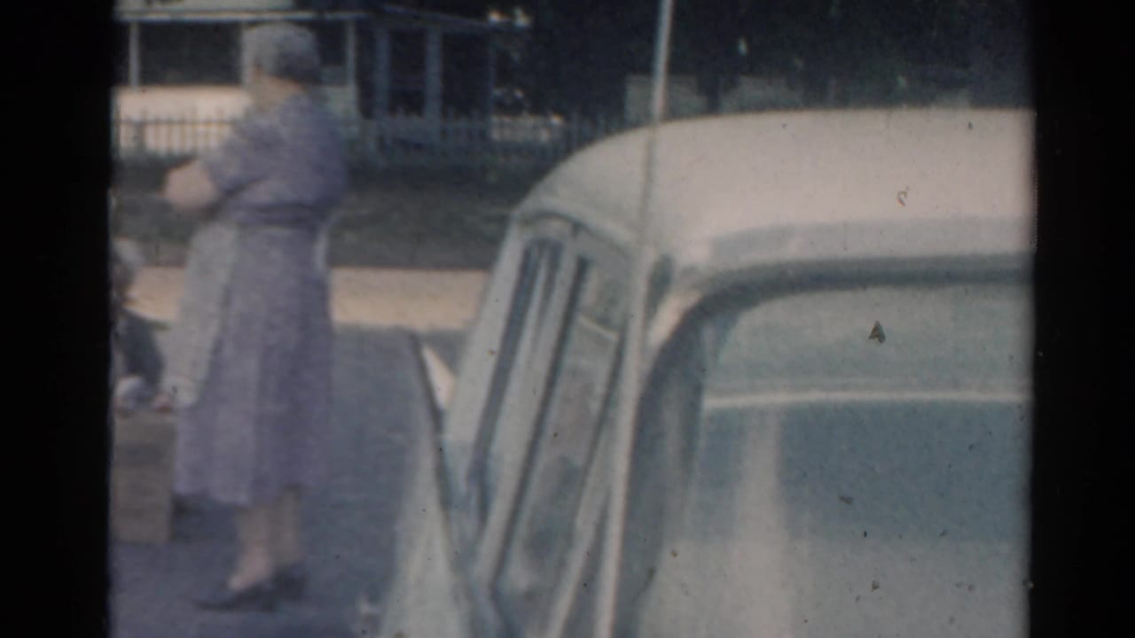 Elderly woman near a parked car on a shaded residential street.