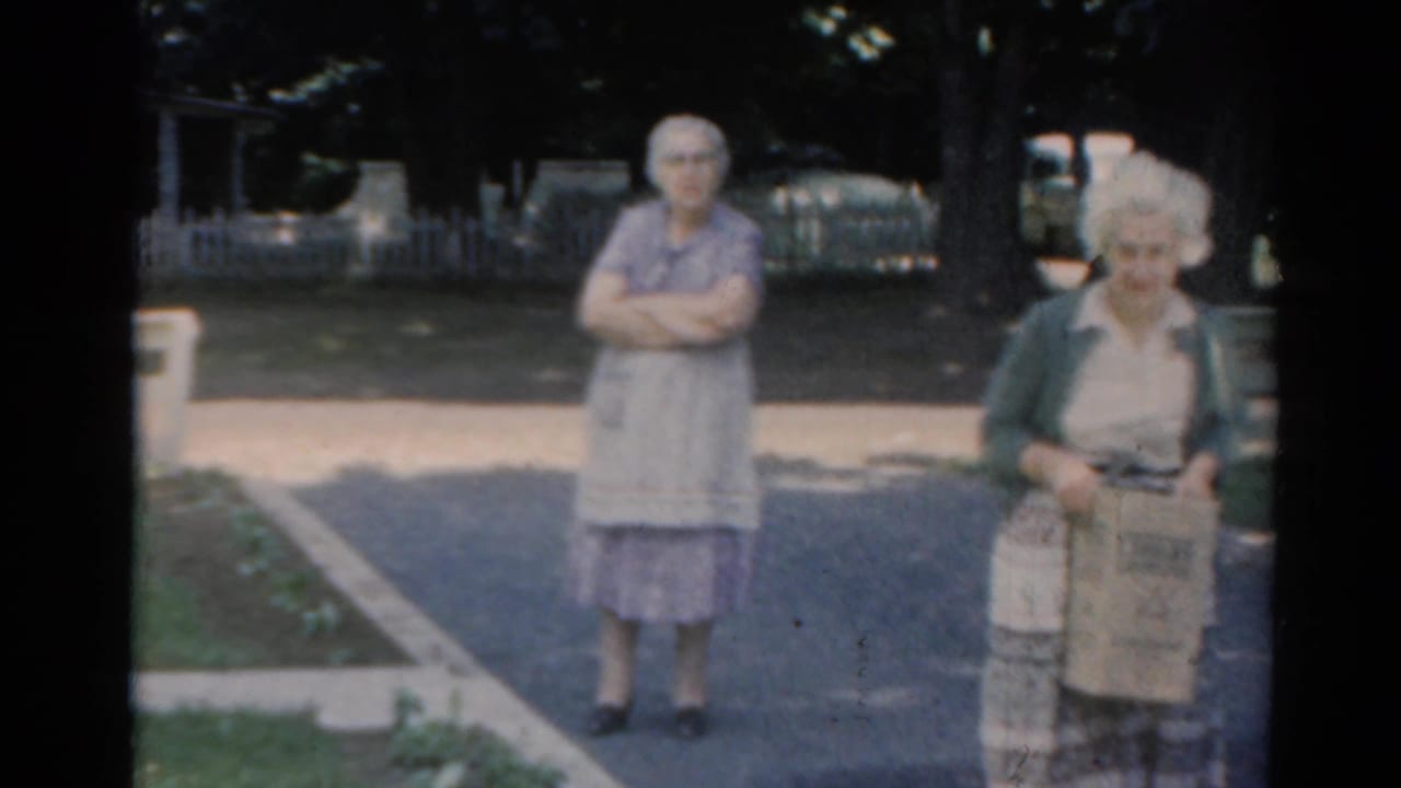 1964 Elkhart, Illinois home-movie frame of two elderly women on a quiet street, with one woman holding a paper bag.