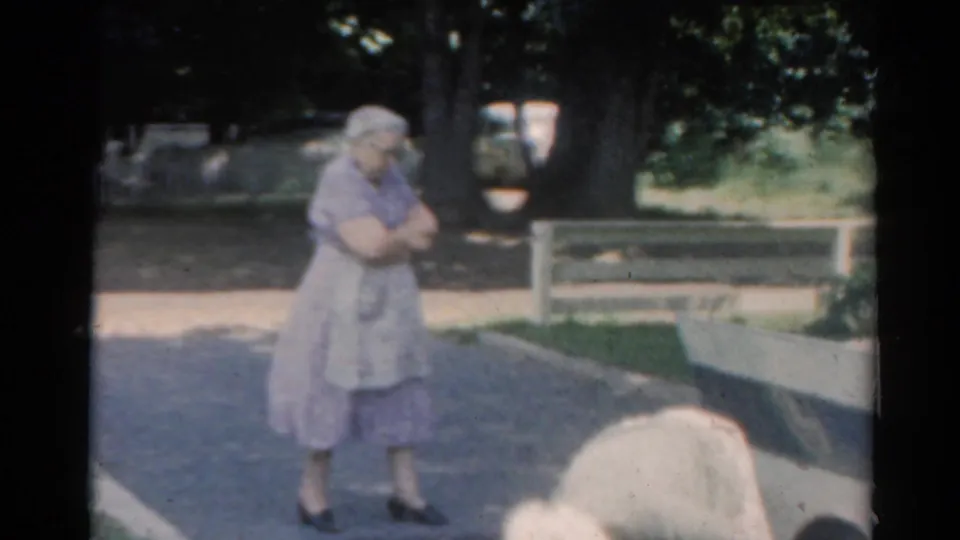 1964 Elkhart, Illinois home-movie frame with an elderly woman standing on the street and a second person partly cropped in front.